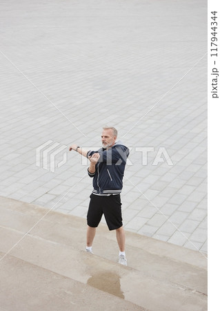 Vertical shot of fit elderly man in sportswear stretching arms standing on concrete steps while getting ready to run jogging against background of tiled square in city park, copy space Vertical shot of fit elderly man in sportswear stretching arms standing on concrete steps while getting ready to run jogging against background of tiled square in city park, copy space 117944344