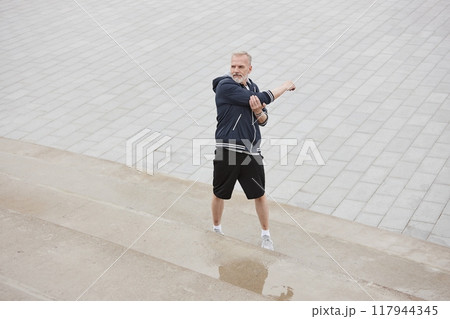 Wide angle shot of fit elderly man in sportswear stretching arms standing on concrete steps while getting ready to run jogging against background of tiled square in city park, copy space 117944345