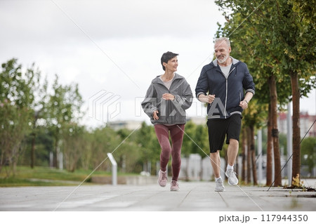 Wide angle shot of fit elderly woman and man in sportswear smiling while jogging together outside along lane of trees working out regularly in morning park, copy space Wide angle shot of fit elderly woman and man in sportswear smiling while jogging together outside along lane of trees working out regularly in morning park, copy space 117944350