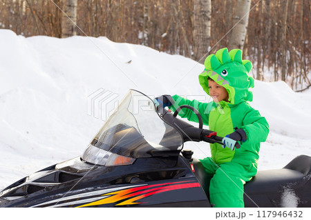 Boy is on a snowmobile in a snowy park in dragon-like outwear. Boy is on a snowmobile in a snowy park in dragon-like outwear. 117946432