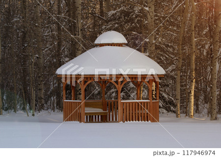 Rustic scene of the wooden cedar brown gazebo in the winter forest covered with snow, warm sunset light. Rustic scene of the wooden cedar brown gazebo in the winter forest covered with snow, warm sunset light. 117946974