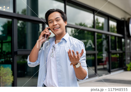 A handsome Asian man is talking on the phone while walking along the building. 117948555
