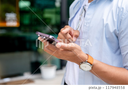 A cropped image of an Asian man in casual clothes is using his smartphone while walking in the city. A cropped image of an Asian man in casual clothes is using his smartphone while walking in the city. 117948558