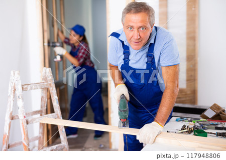 Focused middle-aged man in a blueuniform using a jigsaw machine on wooden plank at home Focused middle-aged man in a blueuniform using a jigsaw machine on wooden plank at home 117948806