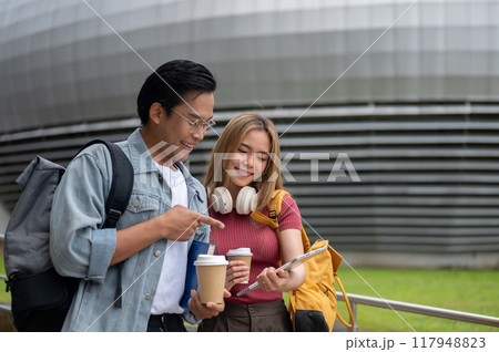 A female college student is showing something on her tablet to her friend while walking together. A female college student is showing something on her tablet to her friend while walking together. 117948823