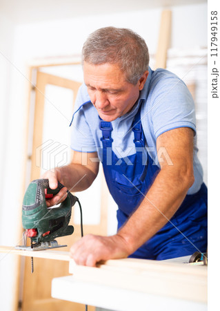 Experienced male worker cuts board with an electric jigsaw in room being renovated Experienced male worker cuts board with an electric jigsaw in room being renovated 117949158