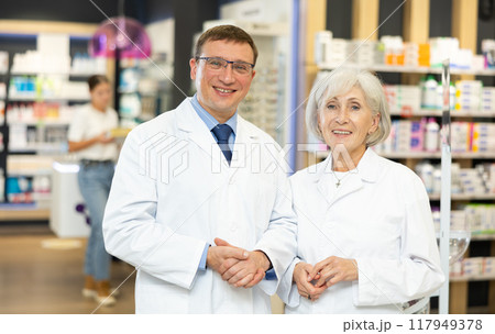 Smiling man and senior female pharmacist workers stand shoulder to shoulder in apotheke Smiling man and senior female pharmacist workers stand shoulder to shoulder in apotheke 117949378