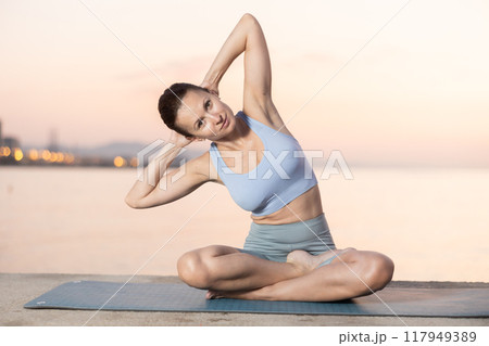 Near sea during sunset, girl performs variations of padmasana with body tilt 117949389