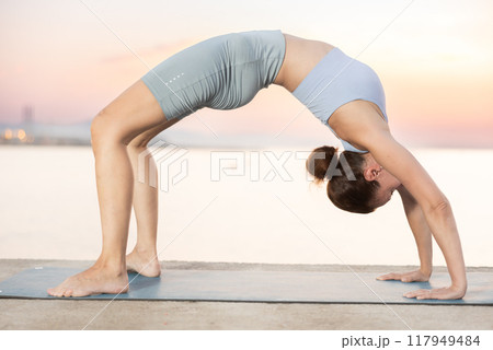 Near sea during sunset, girl performs Ardha chakrasana 117949484