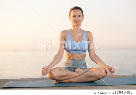 Happy slim woman making yoga meditation in lotus pose on beach at sunrise 117949495