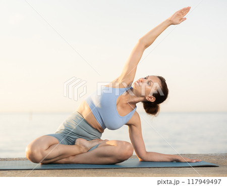 Happy young experienced woman fitness teacher performs yoga parshvottanasana sitting on beach at morning Happy young experienced woman fitness teacher performs yoga parshvottanasana sitting on beach at morning 117949497