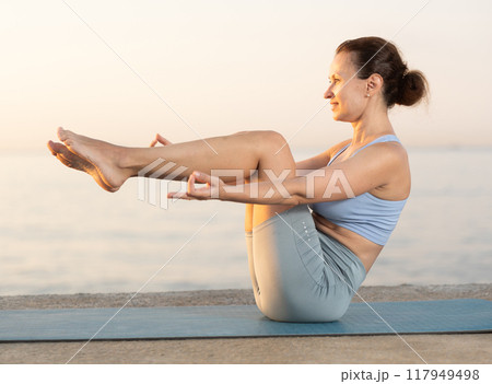 Diligent young woman trainer practicing upward boat pose of yoga, ubhai padangushthasan on mat on seashore at sunset. Philosophy of active lifestyle 117949498
