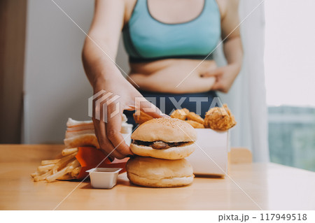 To eat or not. Cheerless plump woman looking at the burger while thinking to eat it or not To eat or not. Cheerless plump woman looking at the burger while thinking to eat it or not 117949518