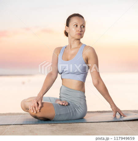 Calm european young female yoga trainer practicing Half Lord of fishes pose or Ardha Matsyendrasana during yoga training on seashore at sunrise Calm european young female yoga trainer practicing Half Lord of fishes pose or Ardha Matsyendrasana during yoga training on seashore at sunrise 117949659