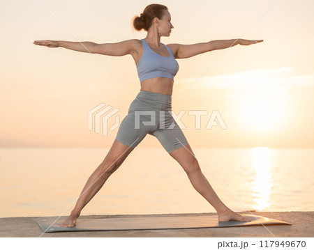 Active experienced woman teacher performs yoga asana on mat on seashore at sunset. Concept of healthy lifestyle 117949670