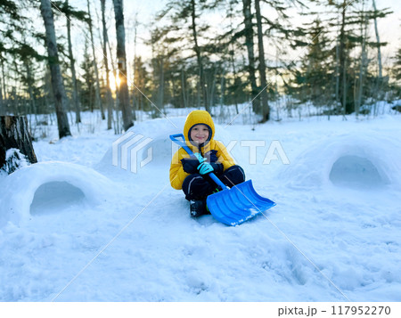 A boy in snowsuit is sitting on the snow in winter yard holding a shovel near built three little igloos at sunset time. A boy in snowsuit is sitting on the snow in winter yard holding a shovel near built three little igloos at sunset time. 117952270