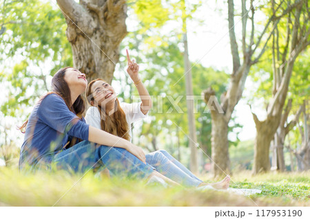 Two Asian women are sitting and relaxing together on the grass in the public park. The two Asian women, both looking good, are sitting and picnicking together on the grass in the public park. 117953190