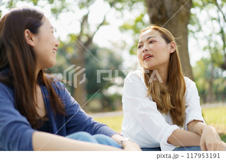 Two Asian women are sitting and relaxing together on the grass in the public park. The two Asian women, both looking good, are sitting and picnicking together on the grass in the public park. 117953191