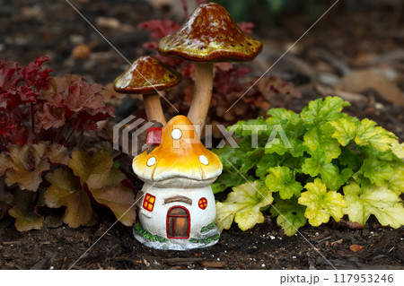 Decorative garden miniature ceramic figurine of a fairy house and mushrooms are among red and green heuchera plants in the flower bed. Decorative garden miniature ceramic figurine of a fairy house and mushrooms are among red and green heuchera plants in the flower bed. 117953246