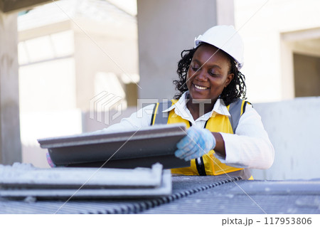 An African American female contractor is examining rooftop tiles at the construction site. The female foreman is inspecting the construction materials. An African American female contractor is examining rooftop tiles at the construction site. The female foreman is inspecting the construction materials. 117953806