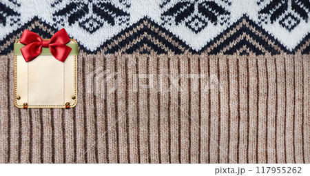 Horizontal Christmas background with knitted fabric texture and leather tag with bow. Wool sweater texture of brown and white color with geometric ornament and blank label. Copy space for text Horizontal Christmas background with knitted fabric texture and leather tag with bow. Wool sweater texture of brown and white color with geometric ornament and blank label. Copy space for text 117955262
