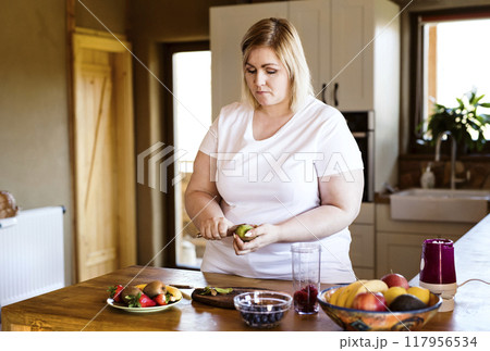 Overweight woman in the kitchen, making fresh smoothie. Choosing healthy food, cutting fruit on cutting board. Overweight woman in the kitchen, making fresh smoothie. Choosing healthy food, cutting fruit on cutting board. 117956534