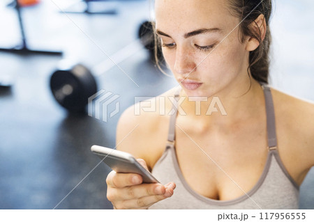 Young fitness girl checking her performance on smatphone after hard workout in gym. Young fitness girl checking her performance on smatphone after hard workout in gym. 117956555
