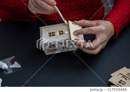 Unrecognizable woman in red sweater painting craft wooden houses for conscientious Merry Christmas decor. Sustainable celebration minimal responsible DIY holiday project. Step by step instruction 117959486