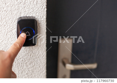 Pressing Black Doorbell with Blue Ring on White Wall 117960730