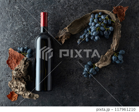 Bottle of red wine, blue grapes, and old vine snags on a black stone background. 117961210