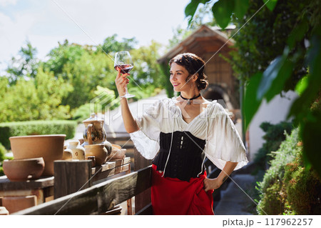 Woman in traditional attire admires glass of red wine, standing on rustic balcony surrounded by pottery, basking in warm glow of sunny day. Concept of vintage, art. 117962257