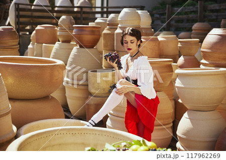 Grapes in hand, lady in thoughtful pose, surrounded by intricate designs of pottery, embodying essence of harvest. Concept of vintage, art. Grapes in hand, lady in thoughtful pose, surrounded by intricate designs of pottery, embodying essence of harvest. Concept of vintage, art. 117962269
