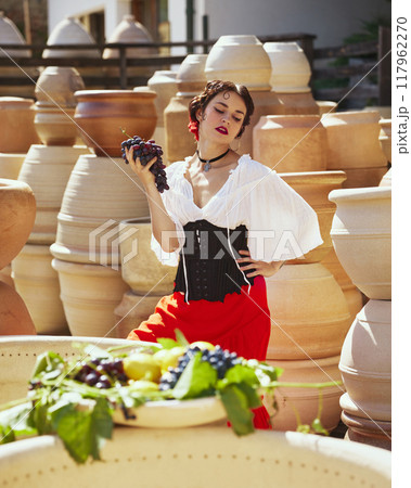 Holding cluster of grapes, woman exudes elegance, her traditional attire blending perfectly with earthy tones of pottery around her. Concept of vintage, art. 117962270