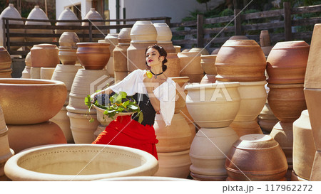 Grapes in hand, woman strikes thoughtful pose, surrounded by intricate designs of pottery, embodying essence of harvest. Concept of tourism and art. Grapes in hand, woman strikes thoughtful pose, surrounded by intricate designs of pottery, embodying essence of harvest. Concept of tourism and art. 117962272