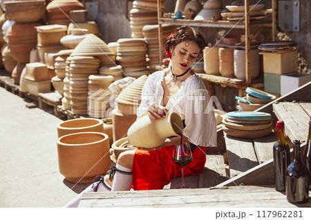 Spanish woman, dressed traditional outfit, elegant girl, Seated amidst large clay pots, she gracefully pours wine from earthenware jug into glass. Concept of tourism and art. Spanish woman, dressed traditional outfit, elegant girl, Seated amidst large clay pots, she gracefully pours wine from earthenware jug into glass. Concept of tourism and art. 117962281