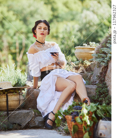 Young woman in white dress, rests with glass of wine, her surroundings celebration of Mediterranean harvest and beauty of countryside. Concept of travelling. Young woman in white dress, rests with glass of wine, her surroundings celebration of Mediterranean harvest and beauty of countryside. Concept of travelling. 117962327