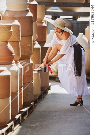 Young Mediterranean woman ties her shoe, surrounded by clay pots, embodying serene blend of autumn's natural beauty and rich cultural history. Concept of harvesting. Young Mediterranean woman ties her shoe, surrounded by clay pots, embodying serene blend of autumn's natural beauty and rich cultural history. Concept of harvesting. 117962344