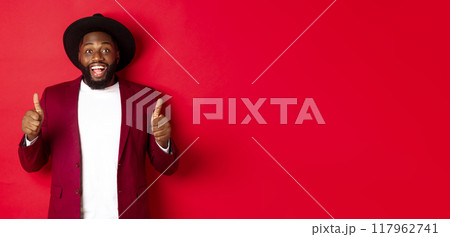 Christmas shopping and people concept. Handsome Black man smiling satisfied, showing thumbs-up, like and agree, approve something, standing against red background Christmas shopping and people concept. Handsome Black man smiling satisfied, showing thumbs-up, like and agree, approve something, standing against red background 117962741