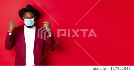 Covid-19, quarantine and holidays concept. Cheerful african american man showing clenched fist and rejoicing of winning, achieve goal, wearing medical mask from coronavirus Covid-19, quarantine and holidays concept. Cheerful african american man showing clenched fist and rejoicing of winning, achieve goal, wearing medical mask from coronavirus 117962848