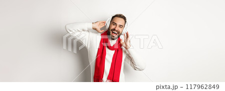 Winter holidays and technology concept. Happy man smiling, listening music in headphones, wearing scarf with sweater, white background 117962849