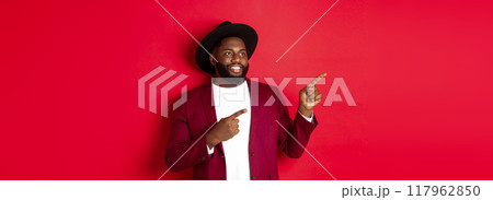 Winter holidays and shopping concept. Happy Black man pointing left and smiling, showing new year promo offer on red background Winter holidays and shopping concept. Happy Black man pointing left and smiling, showing new year promo offer on red background 117962850
