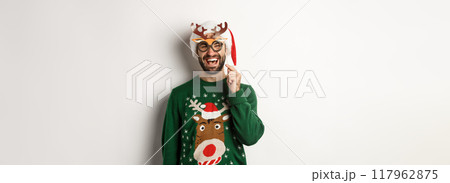 Christmas and holidays concept. Smiling beared man in Santa hat looking happy, holding party mask for New Year, celebrating xmas, standing over white background Christmas and holidays concept. Smiling beared man in Santa hat looking happy, holding party mask for New Year, celebrating xmas, standing over white background 117962875