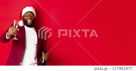 Christmas, party and holidays concept. Handsome Black man in santa hat raising glass of champagne and smiling, saying toast, celebrating New Year, red background Christmas, party and holidays concept. Handsome Black man in santa hat raising glass of champagne and smiling, saying toast, celebrating New Year, red background 117962877