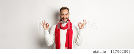 Winter holidays and shopping concept. Excited gay man celebrating christmas, showing okay signs and smiling self-assured, standing over white background Winter holidays and shopping concept. Excited gay man celebrating christmas, showing okay signs and smiling self-assured, standing over white background 117962892