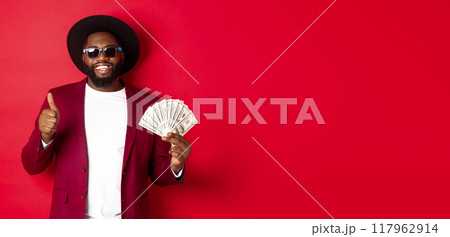 Handsome modern african american man in sunglasses and party clothes, showing thumb up with dollars, earn money and looking satisfied, red background 117962914