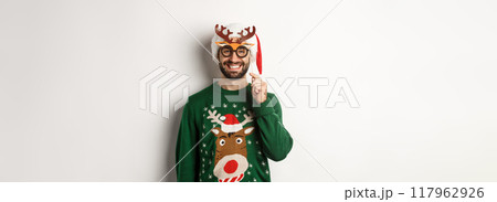 Christmas and holidays concept. Smiling beared man in Santa hat looking happy, holding party mask for New Year, celebrating xmas, standing over white background Christmas and holidays concept. Smiling beared man in Santa hat looking happy, holding party mask for New Year, celebrating xmas, standing over white background 117962926