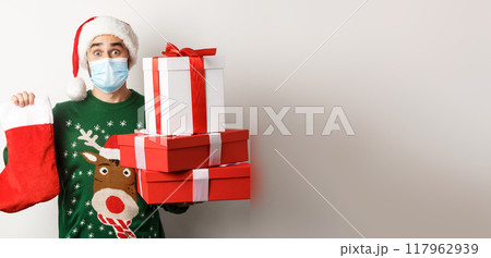 Winter holidays and xmas concept. Happy man in face mask and santa hat bringing gifts, holding Christmas sock and present boxes, standing over white background 117962939