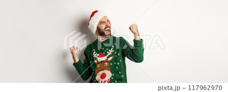 Christmas holidays, celebration and party concept. Happy guy in Santa hat and sweater, making fist pumps and rejoicing, triumphing, standing over white background Christmas holidays, celebration and party concept. Happy guy in Santa hat and sweater, making fist pumps and rejoicing, triumphing, standing over white background 117962970