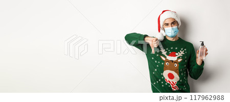 Concept of covid-19 and Christmas holidays. Disappointed man in face mask and santa hat, dislike hand sanitizer, showing thumbs down, standing over white background Concept of covid-19 and Christmas holidays. Disappointed man in face mask and santa hat, dislike hand sanitizer, showing thumbs down, standing over white background 117962988