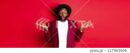 Christmas shopping and people concept. Handsome african american man staring amazed, spread hands sideways, showing promo offers on copy space, red background Christmas shopping and people concept. Handsome african american man staring amazed, spread hands sideways, showing promo offers on copy space, red background 117963009